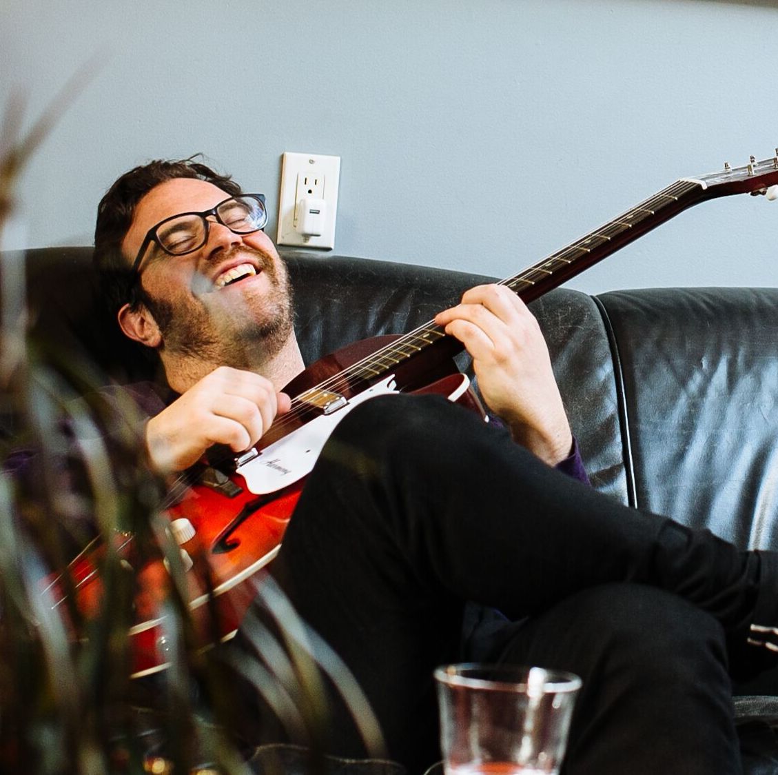 Joel with a guitar, sitting on a couch smiling.